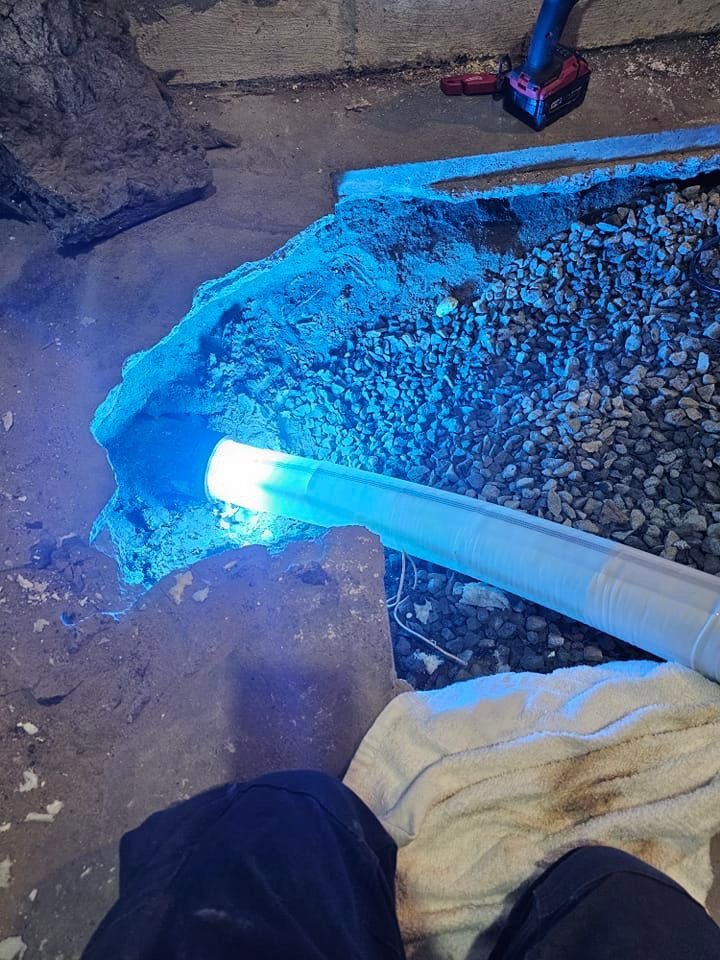 A construction site shows a partially buried pipe emitting a bright blue light, with gravel around it and a towel-covered surface in the foreground. A power tool is visible on the right side, highlighting the contrasting nature of sewer pipe lining vs residential excavation in modern engineering solutions.