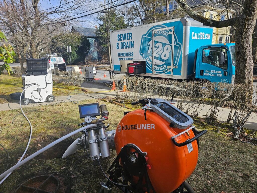A 128 Plumbing truck labeled "Sewer & Drain Trenchless" and equipment including an orange "HouseLiner" device are set up on a residential street, with hoses and cables extending from them.