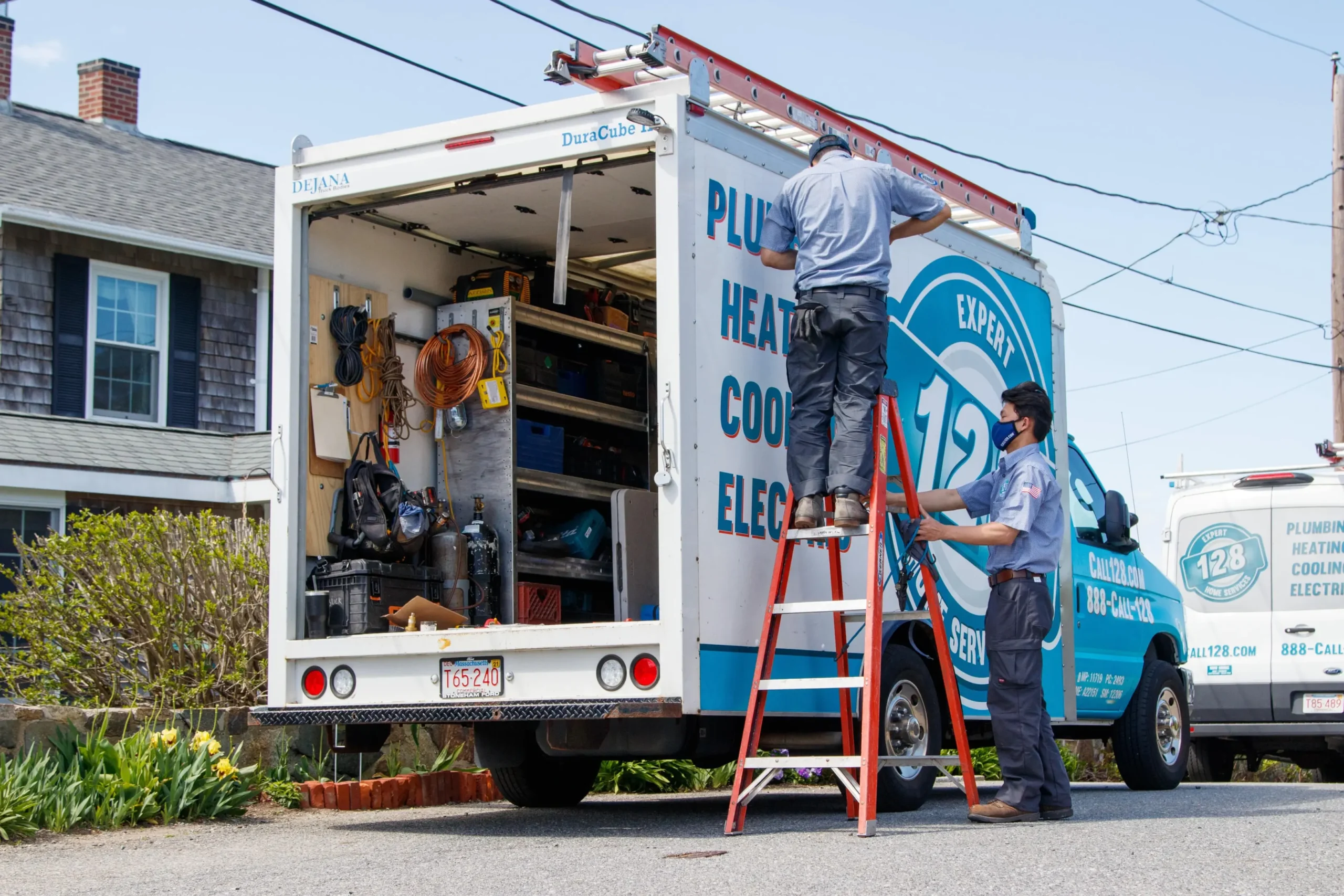 Two workers in uniform organize equipment in the back of a plumbing and heating service truck. One worker stands inside the truck, and the other adjusts a ladder. The truck has various tools and supplies.