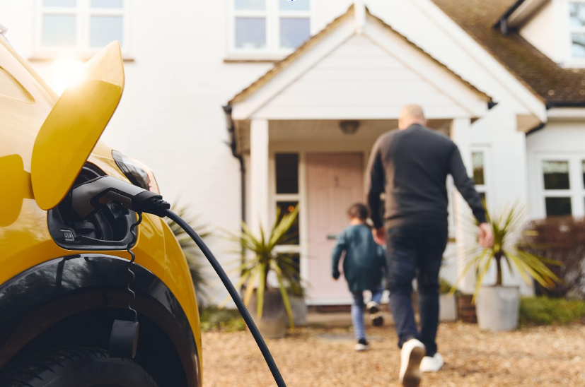 A yellow electric car charges in the foreground as a man and child walk toward a white house with a pink front door.