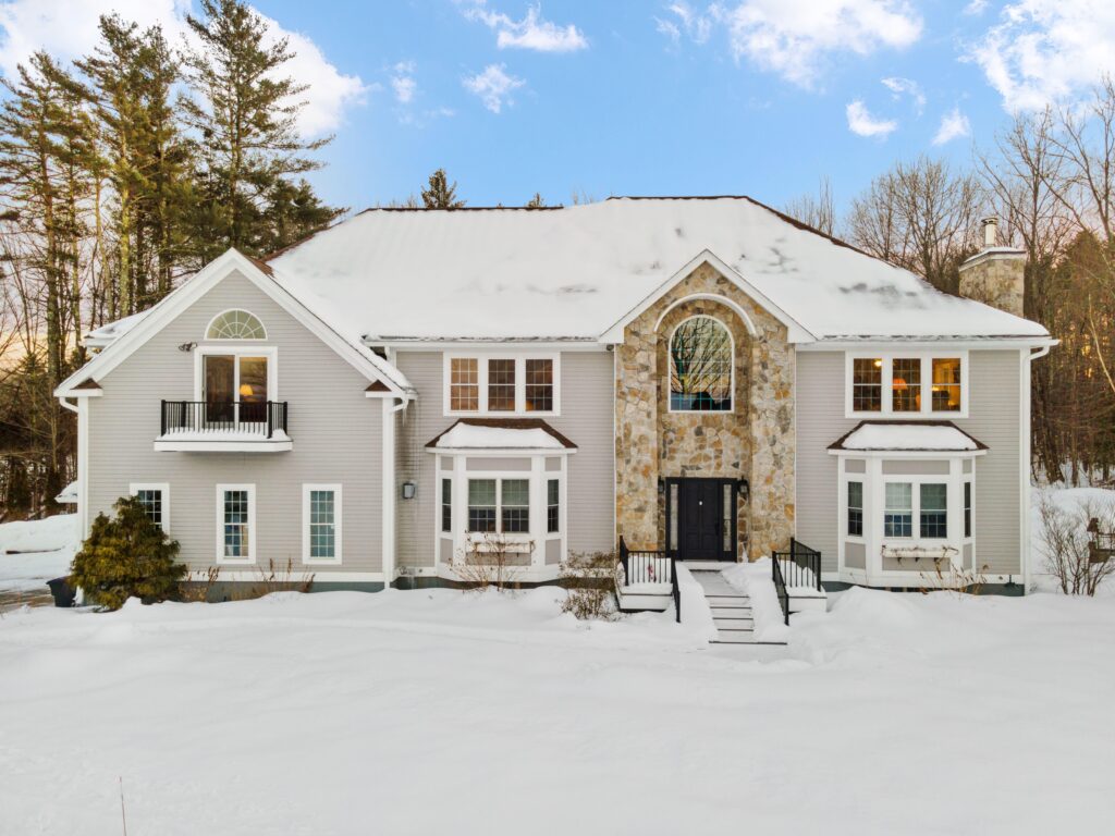 A large two-story house with a stone facade and snow-covered ground, featuring symmetrical windows, dormers, and a central entrance with stairs leading up to a black front door.