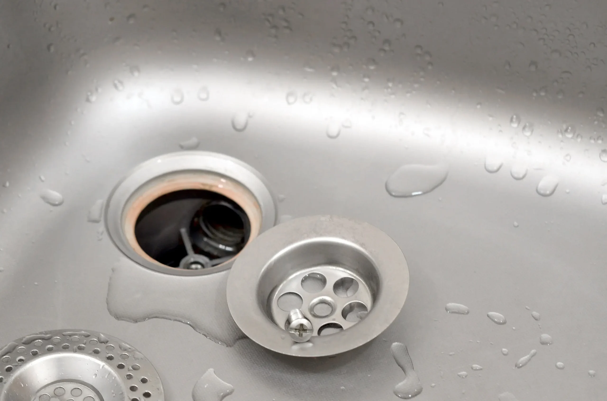 A stainless steel sink with water droplets and two removable drain strainers, one of which is partially dislodged, exposing the drain hole.