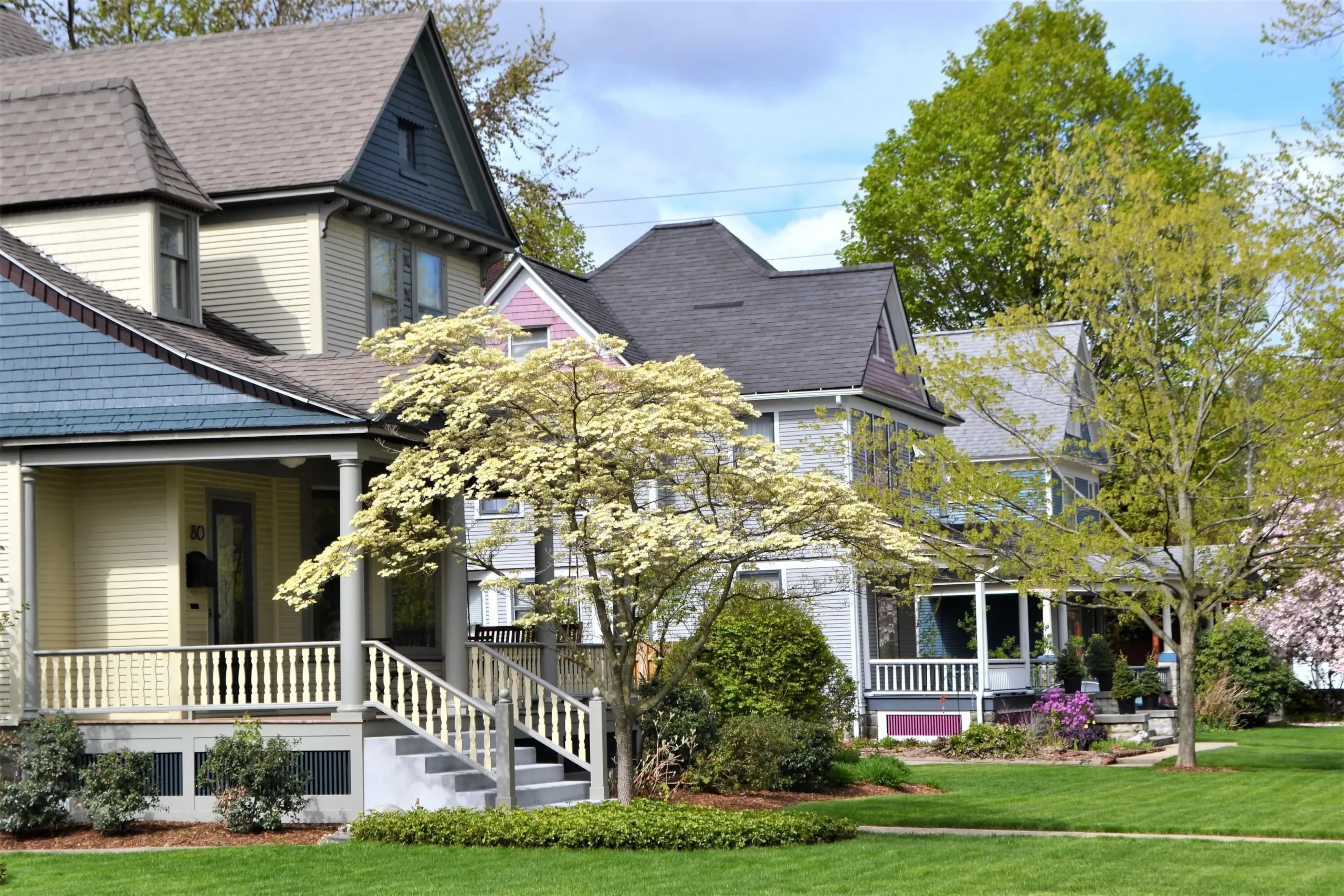 Row of colorful houses with porches, lush green lawns, and blooming trees under a partly cloudy sky.