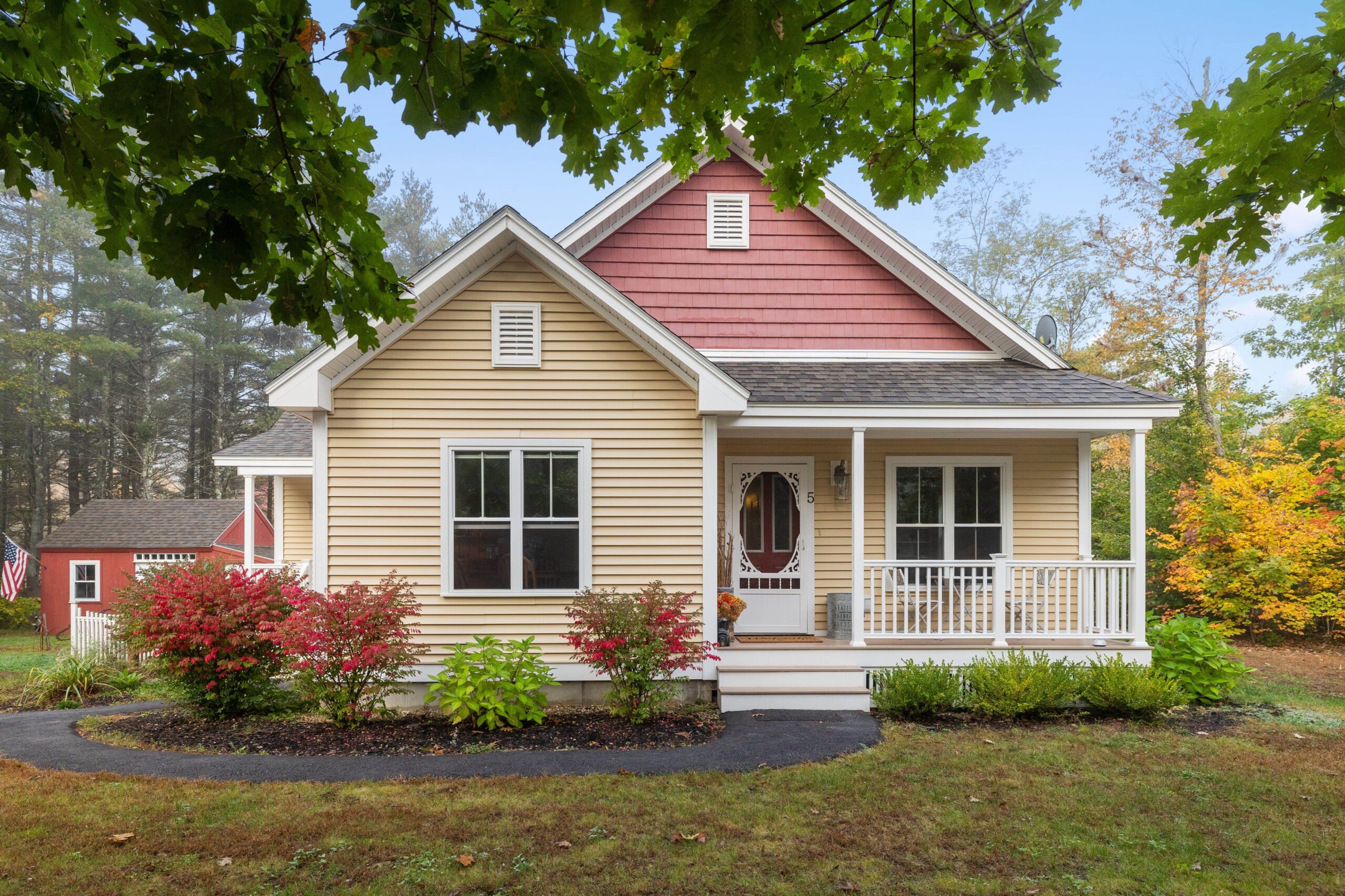 A small beige and red house with a front porch and a white railing, surrounded by a green lawn, shrubs, and trees. A red shed is visible in the background.