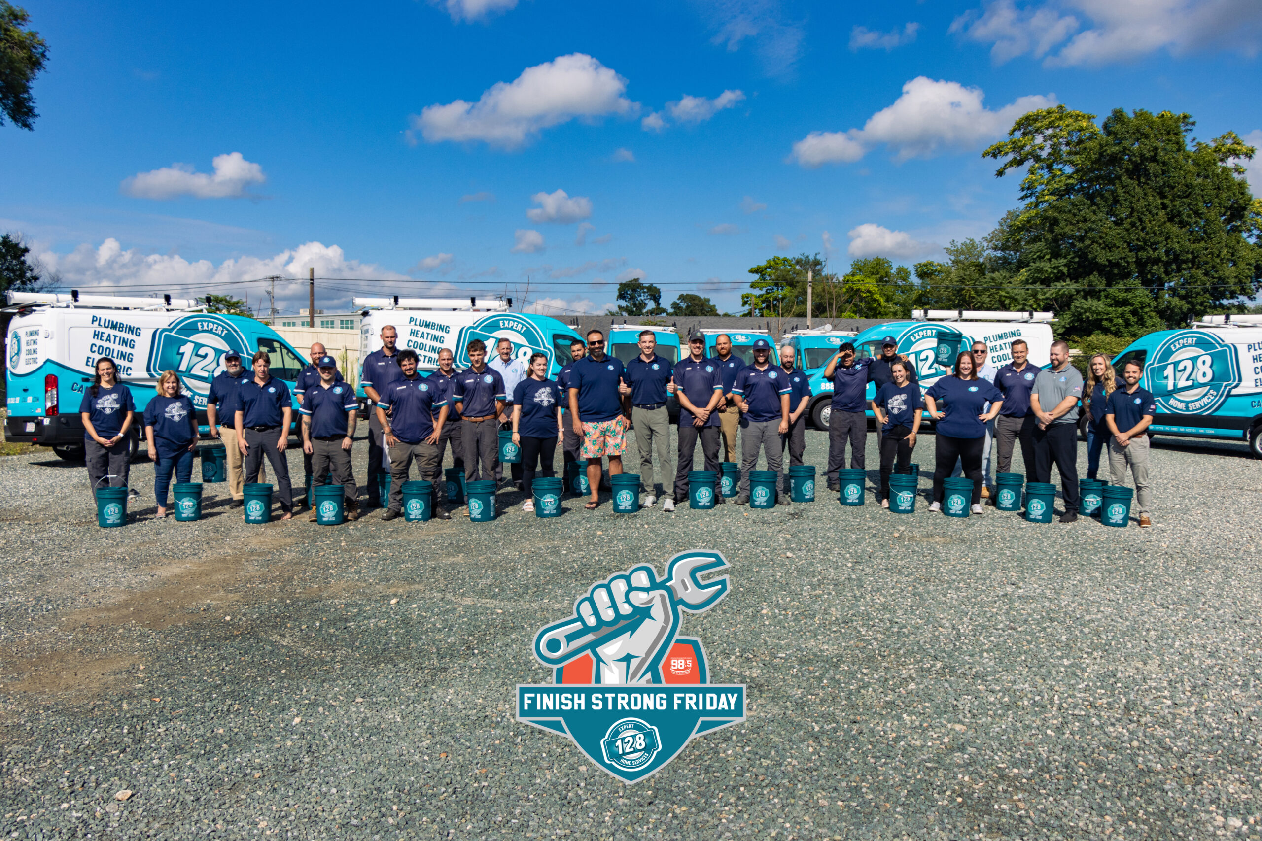 A group of people in uniform stands in front of branded vans on a gravel lot, holding bags with the logo "Finish Strong Friday" and the number 128. The sky is clear with few clouds.