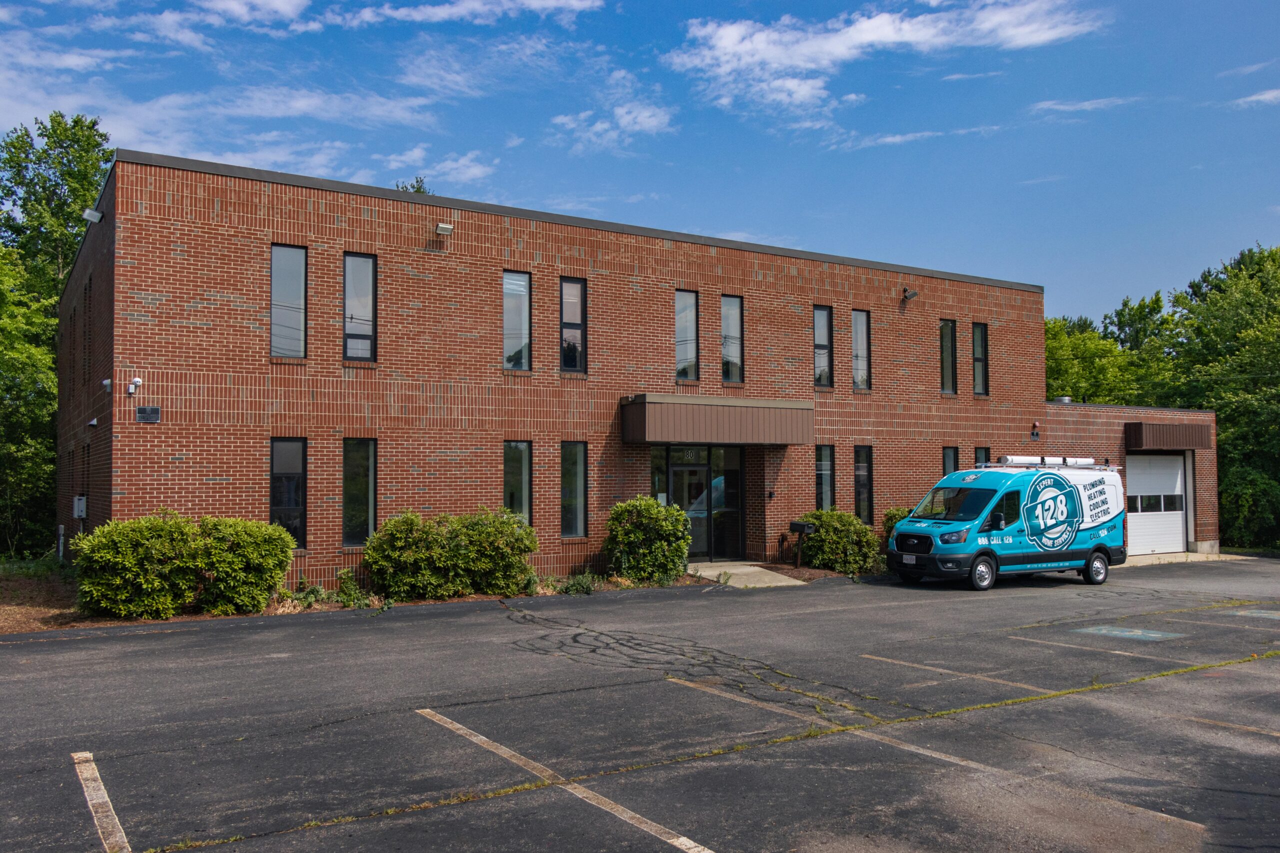 A two-story brick building with several vertical windows stands tall. A blue van labeled "128 Plumbing" is parked in front of the new Chelmsford office on an empty paved lot. Trees are in the background under a clear sky.